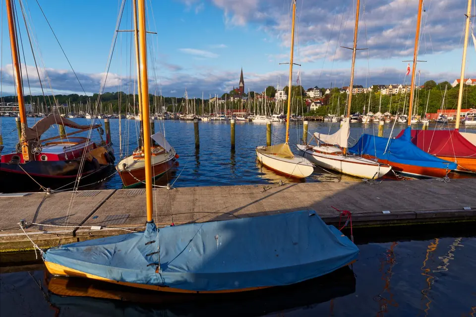 Charmanter Blick auf Segelboote, die im Hafen von Flensburg festgemacht sind, mit der Stadtlandschaft unter sommerlichem Himmel.